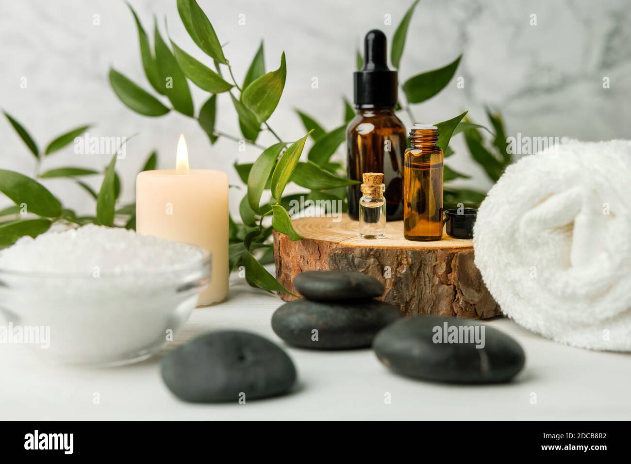 beauty treatment items for spa procedures on white wooden table with green plant. massage stones, essential oils and sea salt with burning candle Stock Photo