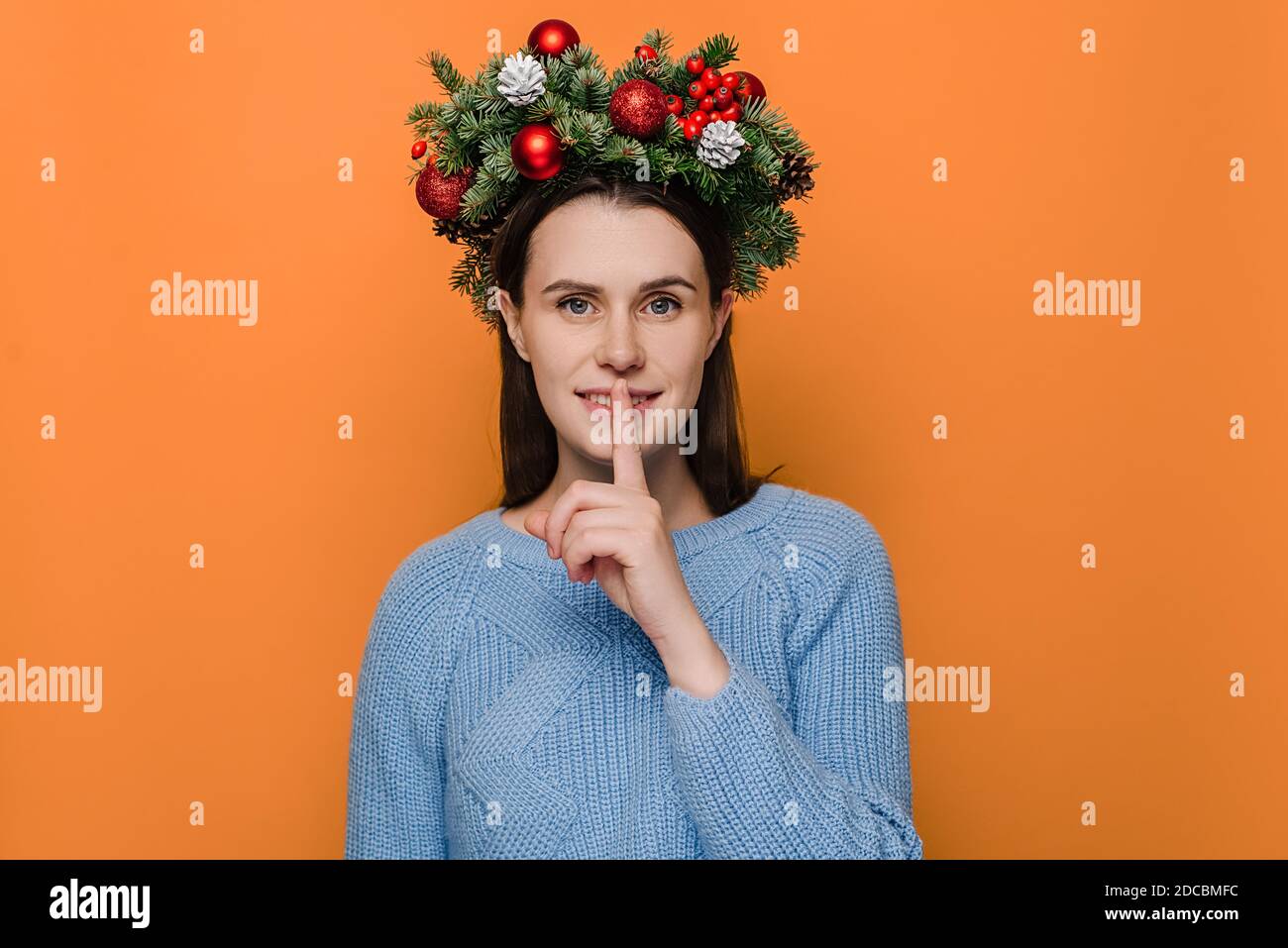 Positive female in handmade wreath, has toothy smile, makes hush gesture, wears winter sweater, poses against orange wall with copy space Stock Photo