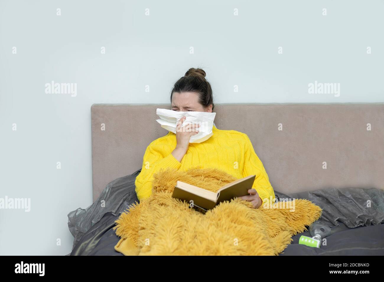 Woman reading book in bed, with paper napkin sneezing, experiences allergy symptoms, caught a cold, stay isolation for self quarantine. Concept home Stock Photo