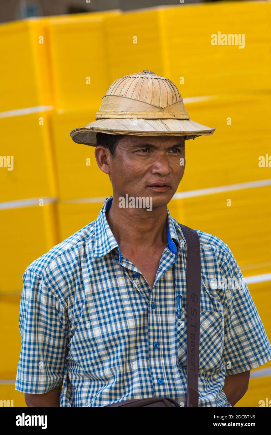 Daily life in Myanmar - man wearing traditional Burmese bamboo hat and shirt set against yellow backdrop at Yangon, Myanmar (Burma), Asia in February Stock Photo