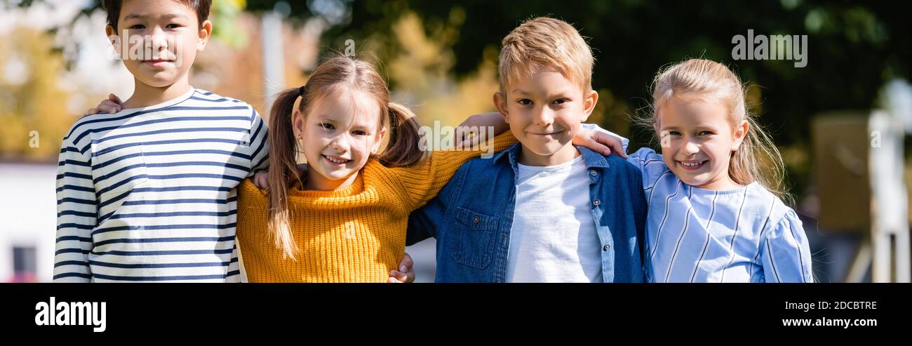 Cheerful multiethnic children hugging while looking at camera outdoors, banner Stock Photo