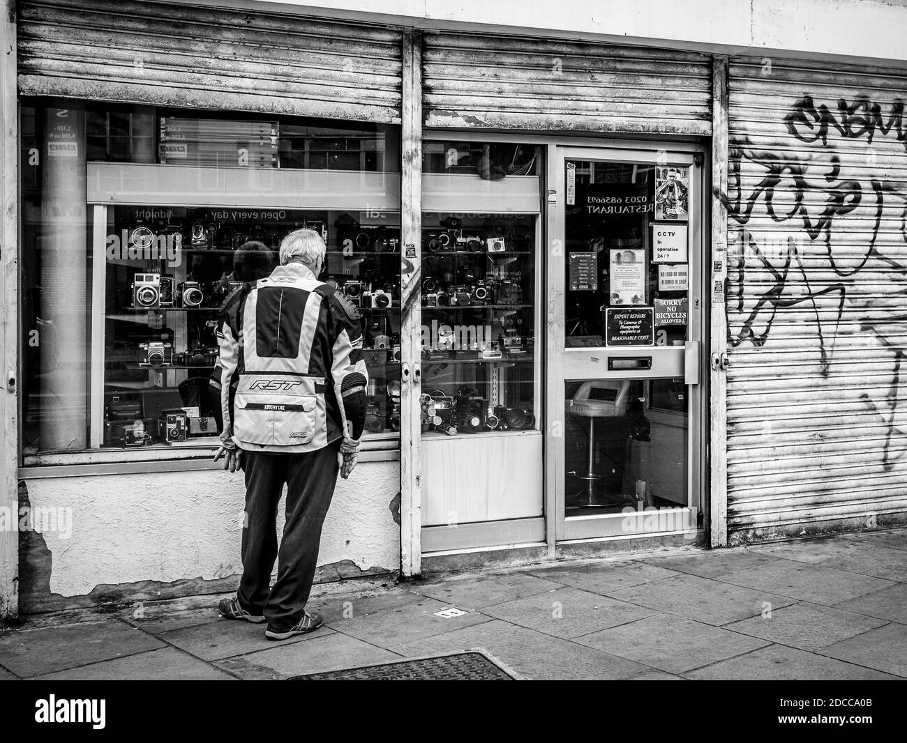 Man window shopping for cameras in Camden Town London with partially shut steel shutters with graffiti on them in a black and white tone, 15 Aug 2020 Stock Photo