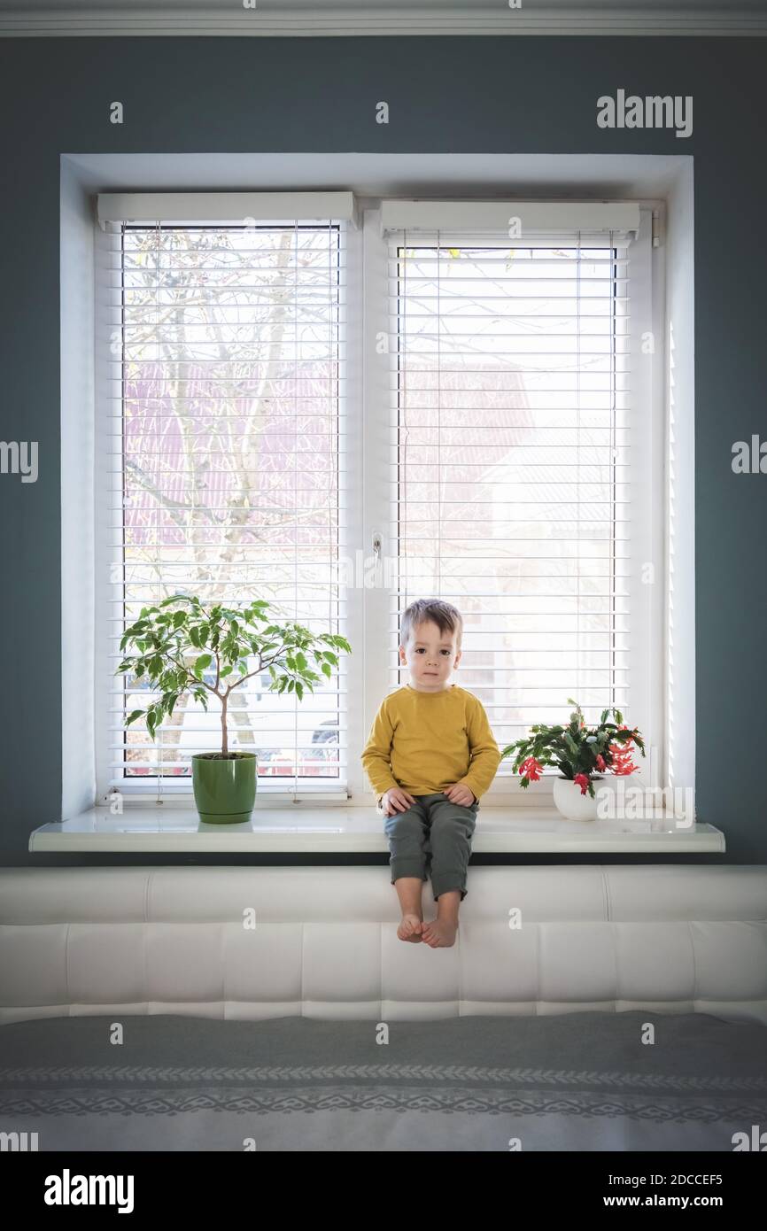Little loneliness kid boy sitting on windowsill near large window in his room. Childhood concept Stock Photo