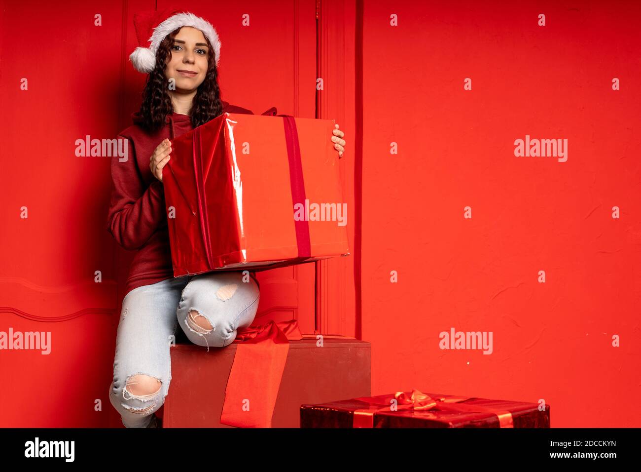 Young attractive woman in Santa Claus hat sits on gifts near red wall. Smiley female sitting on big box of gift. Concept of holidays, gifts and good m Stock Photo