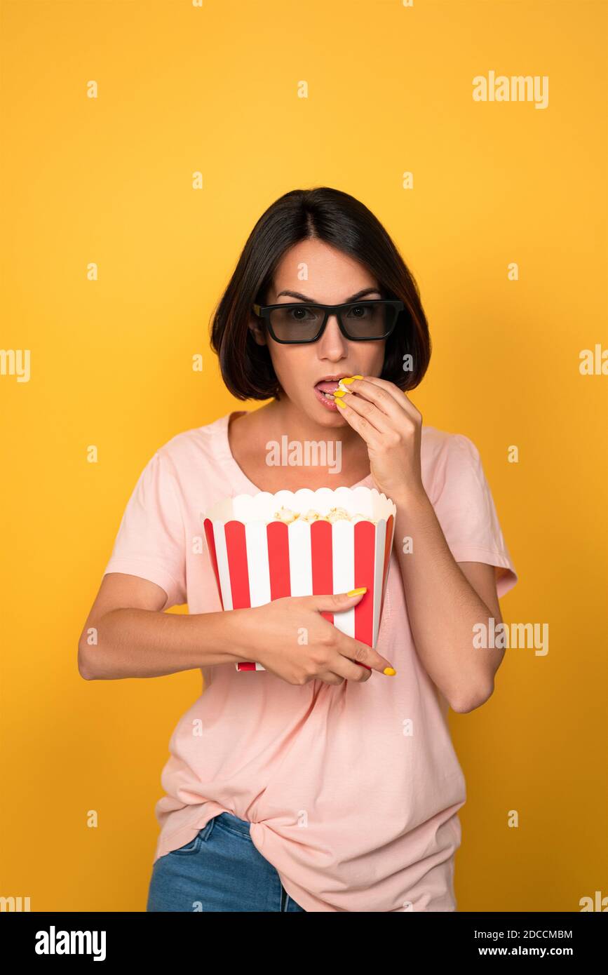 Young brunette woman with bucket of popcorn in stereo glasses. Cinema concept. Isolated on yellow background Stock Photo