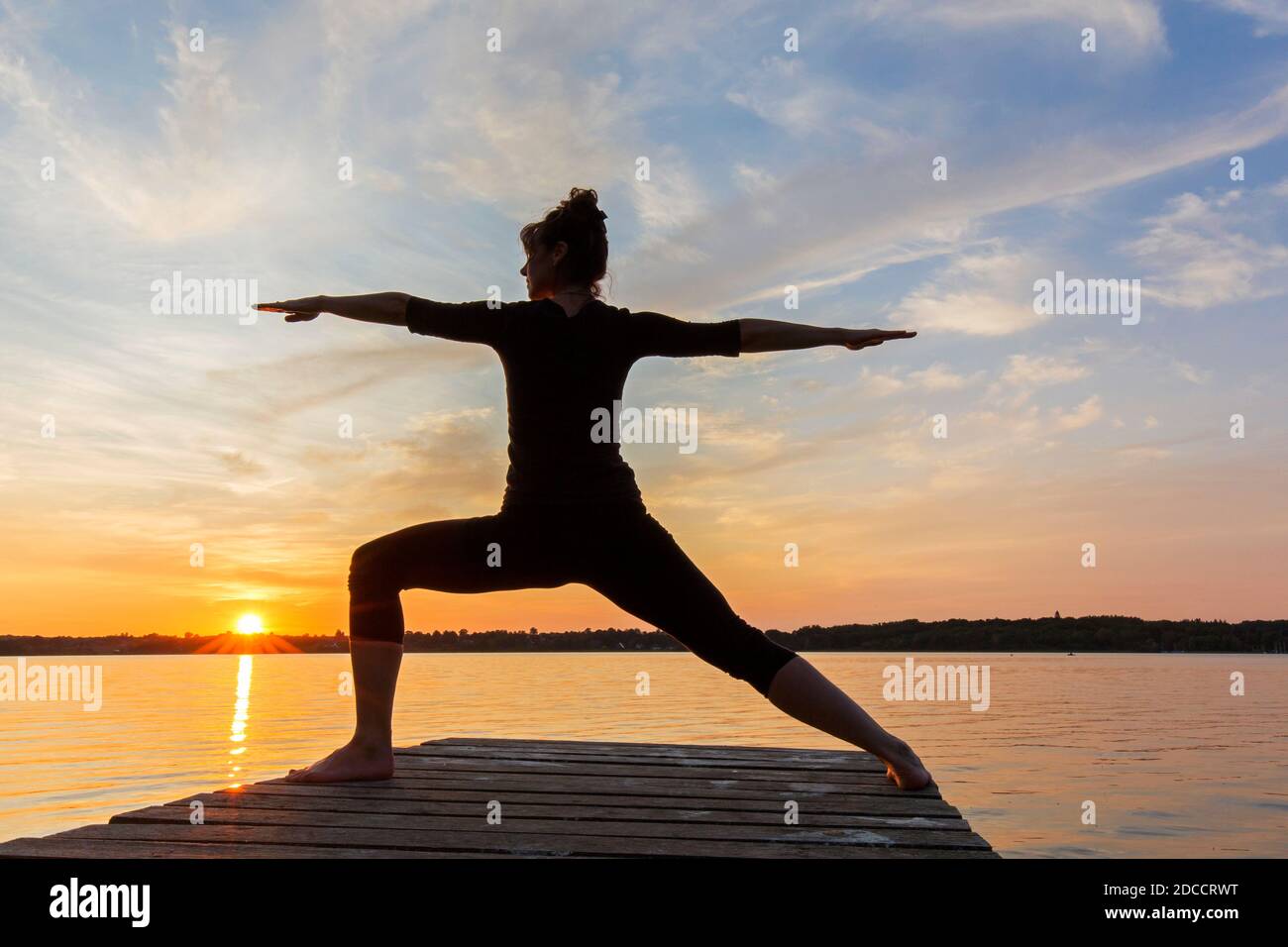 Woman practicing yoga posture Virabhadrasana II / Warrior Pose, lunging standing asana on jetty at lake at sunset Stock Photo