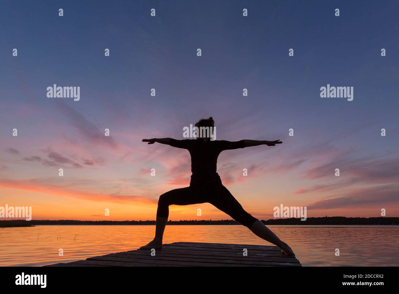 Woman practicing yoga posture Virabhadrasana II / Warrior Pose, lunging standing asana on jetty at lake at sunset Stock Photo