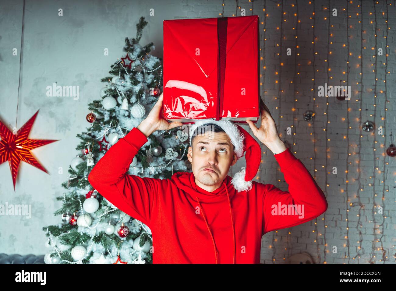 Young handsome man in Santa Claus hat dancing with big gift on background of Christmas tree. Happy guy dances, rejoicing present. Concept of holidays, Stock Photo