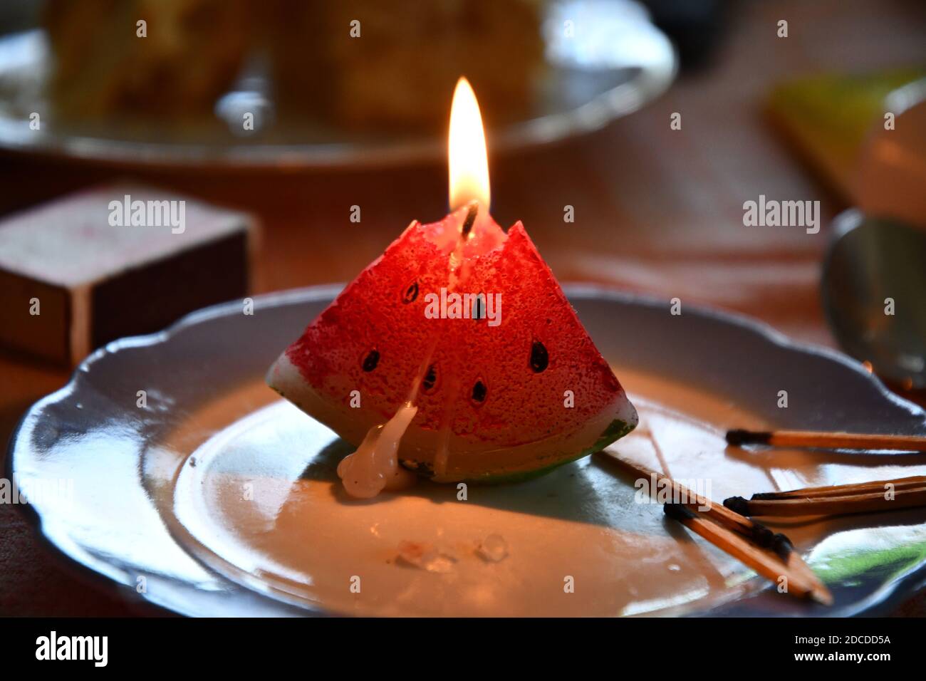 Melted candle in shape of triangle piece of watermelon. Candlelight flame closeup. Burning fire of candle on vintage plate with burnt matches nearby Stock Photo