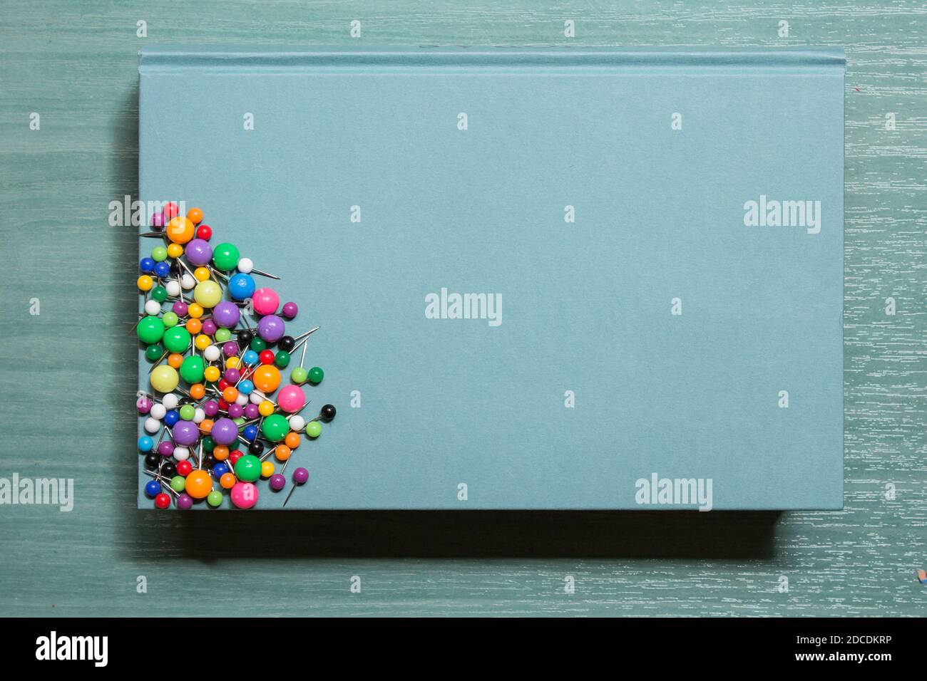 A blue book with push pins of different sizes, shapes, and colors rests on a beautiful wooden table. Stock Photo