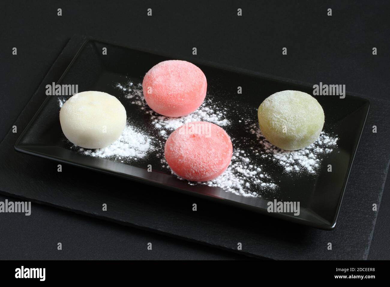 Four types of Japanese dessert mochi - pomegranate with honey, green matcha tea, strawberry, coconut on a black plate on a black table. Closeup Stock Photo