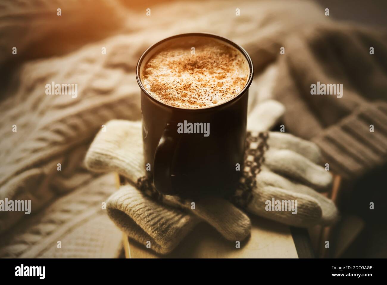 A mug of hot, aroma coffee with cream and cinnamon sprinkles sits on top of a stack of old books and white wool gloves with patterns, and a warm sweat Stock Photo