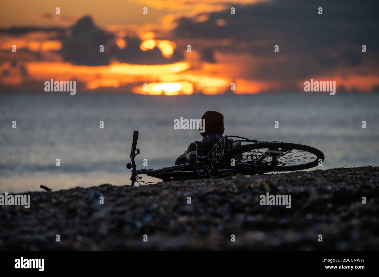 A male cyclist resting on a beach while watching the sunset, illustrating time to sit and think alone. Stock Photo