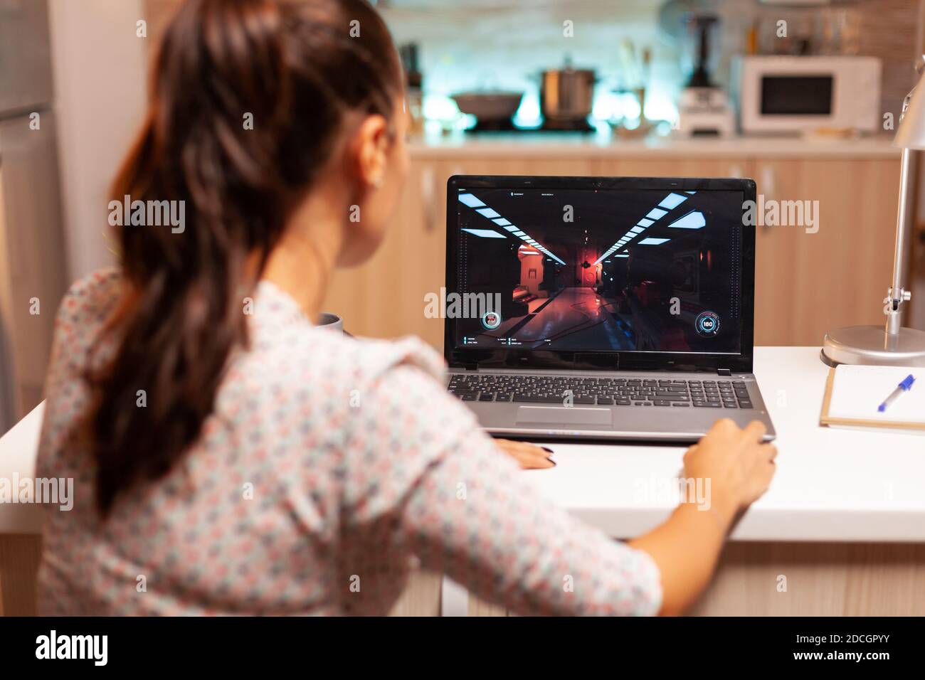 Lady playing games on laptop during night time during night time in home kitchen. Professional gamer playing online videop games on her personal computer. Geek cyber e-sport. Stock Photo