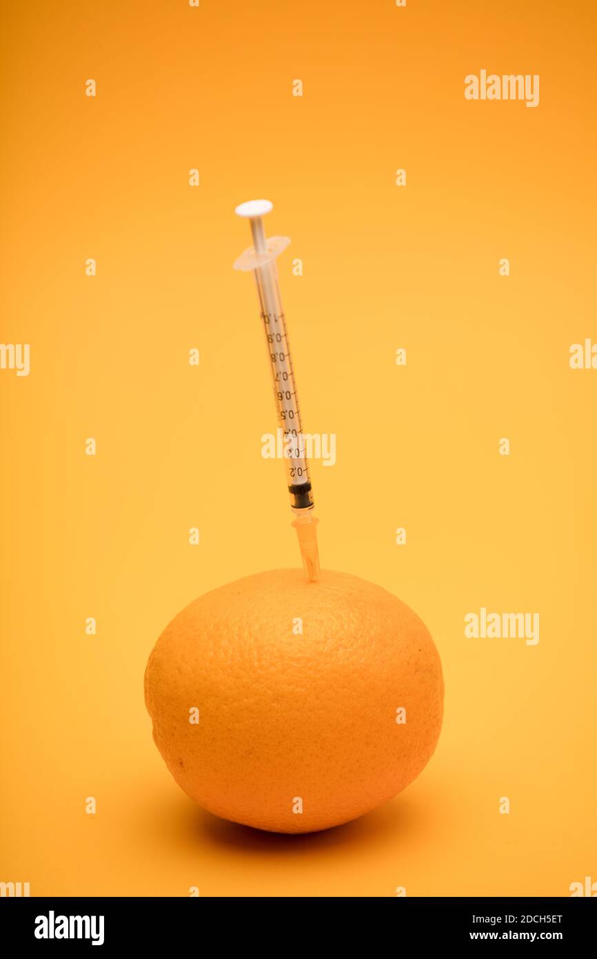 orange with syringe stuck, genetic modification experiment, orange background Stock Photo