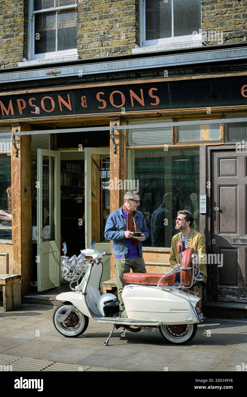 Two young men with lambretta scooter drinking coffee in front of the Climpson & Sons cafe in East London . Stock Photo