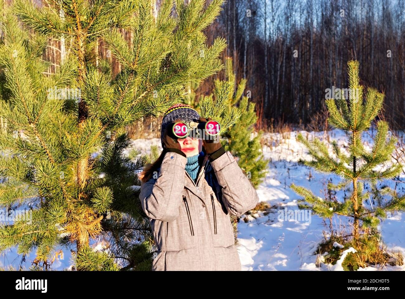 upcoming new year 2021 and christmas concept. Young woman in knitted hat and scarf looking through binoculars with numbers 2021 in eyepieces in winter Stock Photo