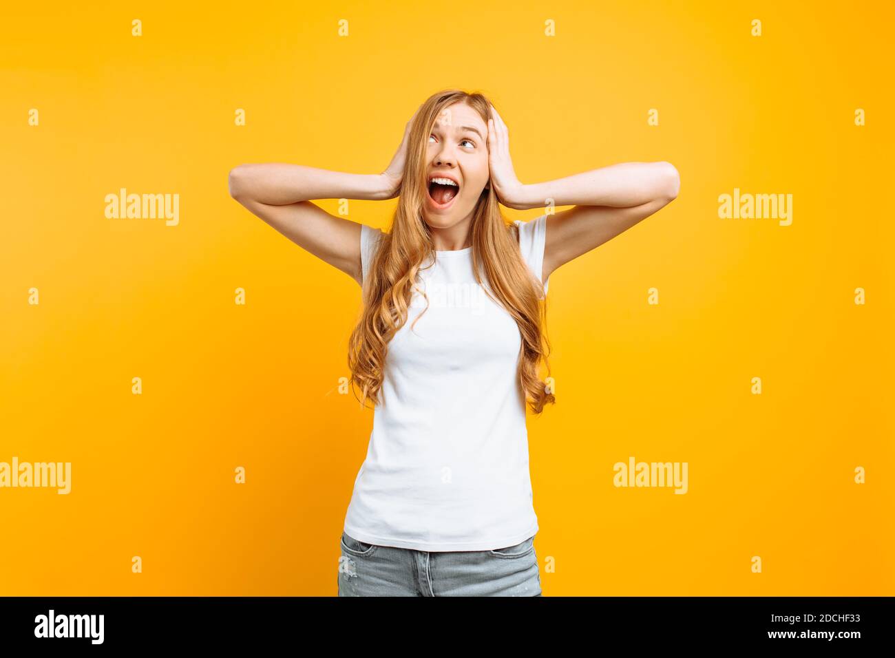 Portrait of an irritated woman in a white T-shirt covering her ears and screaming from a terrible noise, on a yellow background Stock Photo