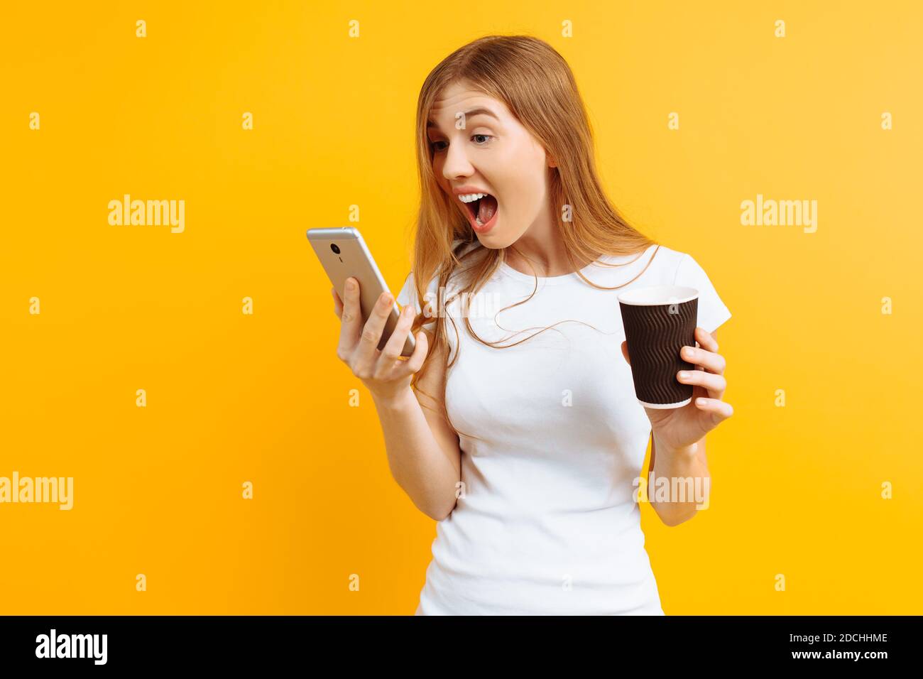 Happy young woman in a white T-shirt, holding a glass of coffee in her hands, and talking on a mobile phone and shouting against a yellow background Stock Photo