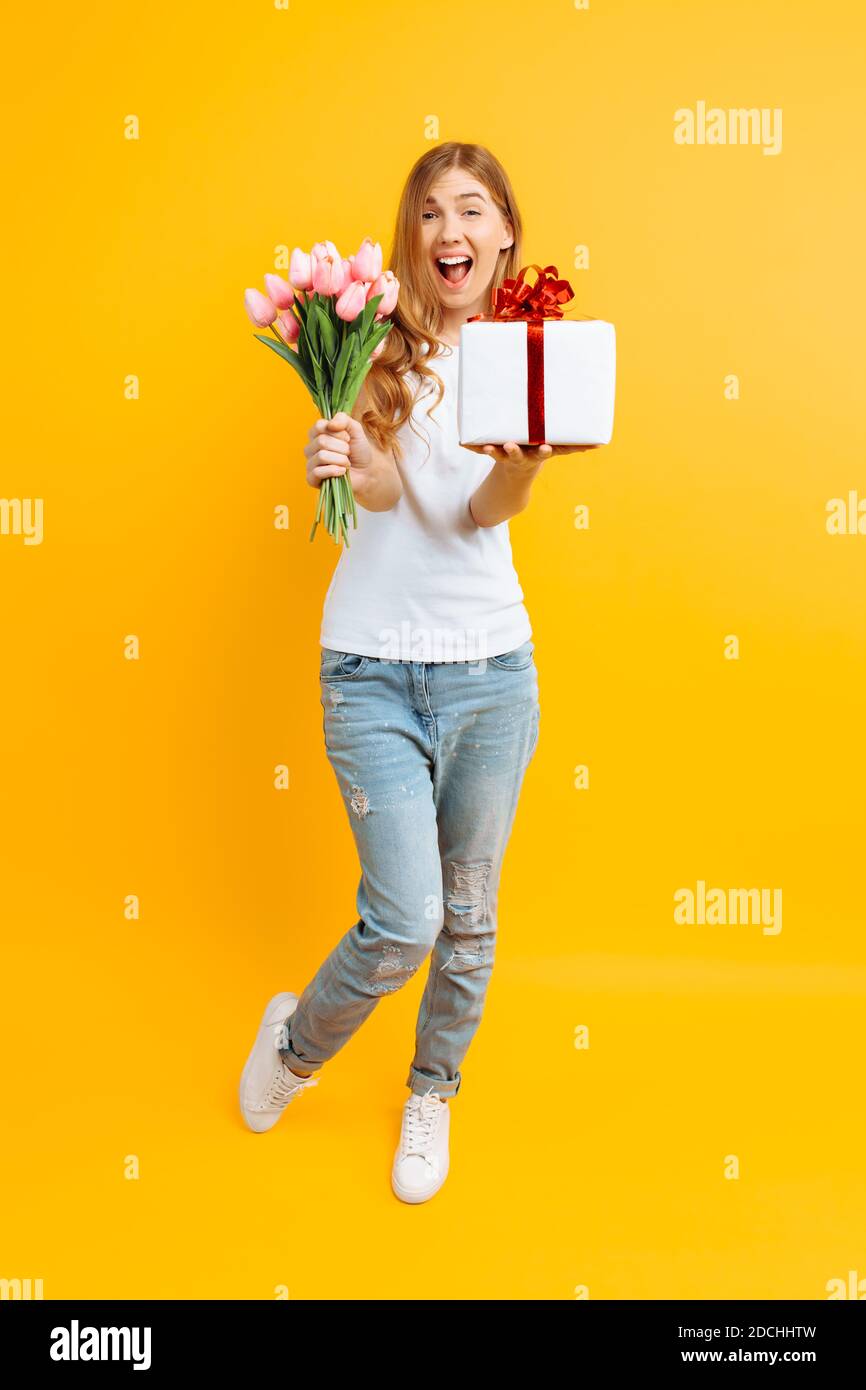 Full length, enthusiastic girl screaming with happiness in a white T-shirt with a bouquet of beautiful flowers and a gift box on a yellow background. Stock Photo