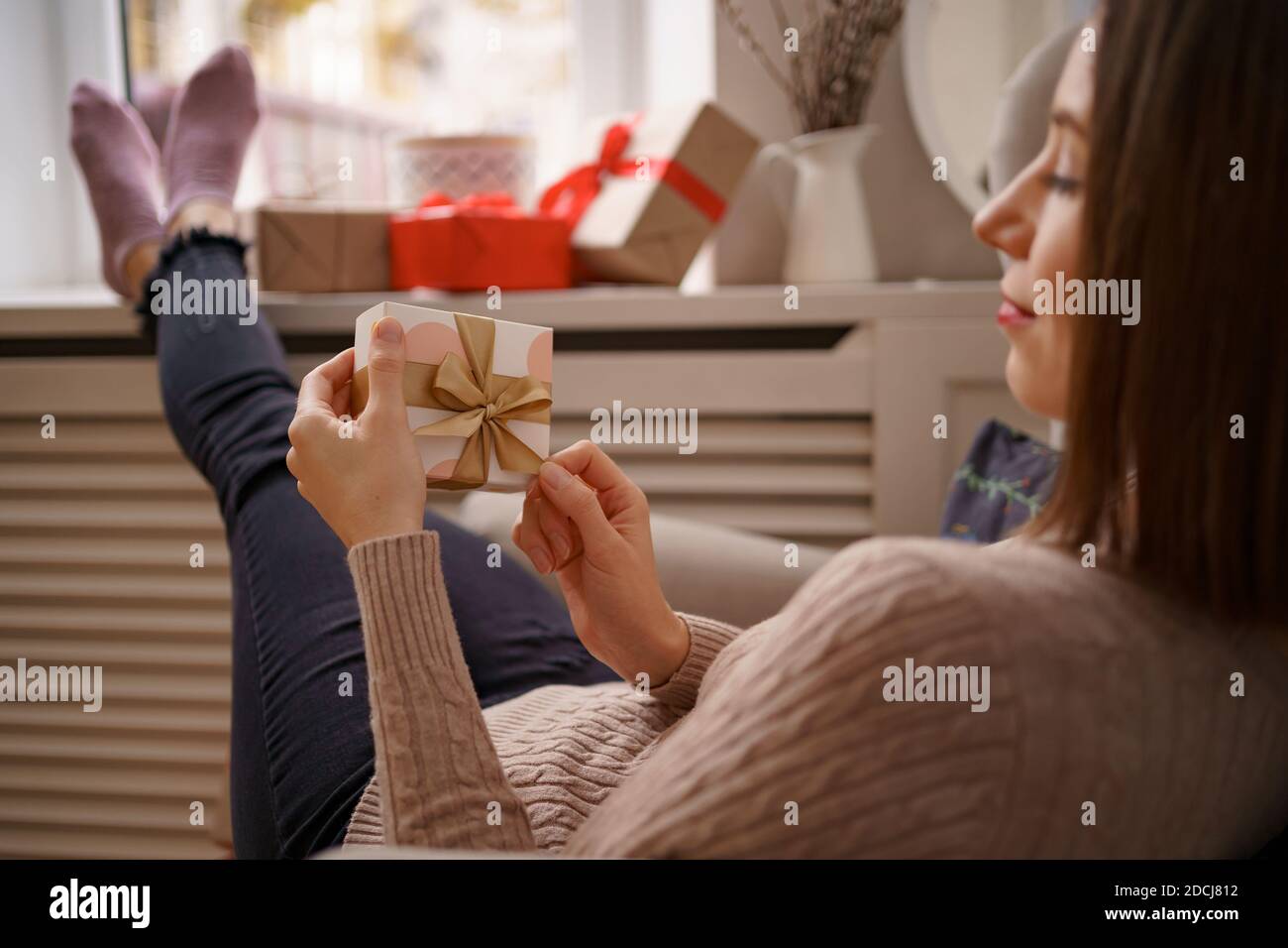 PRETTY young woman while sitting in a comfortable armchair holding a gift box surrounded by gifts at home. Ghe guesses what's inside Stock Photo