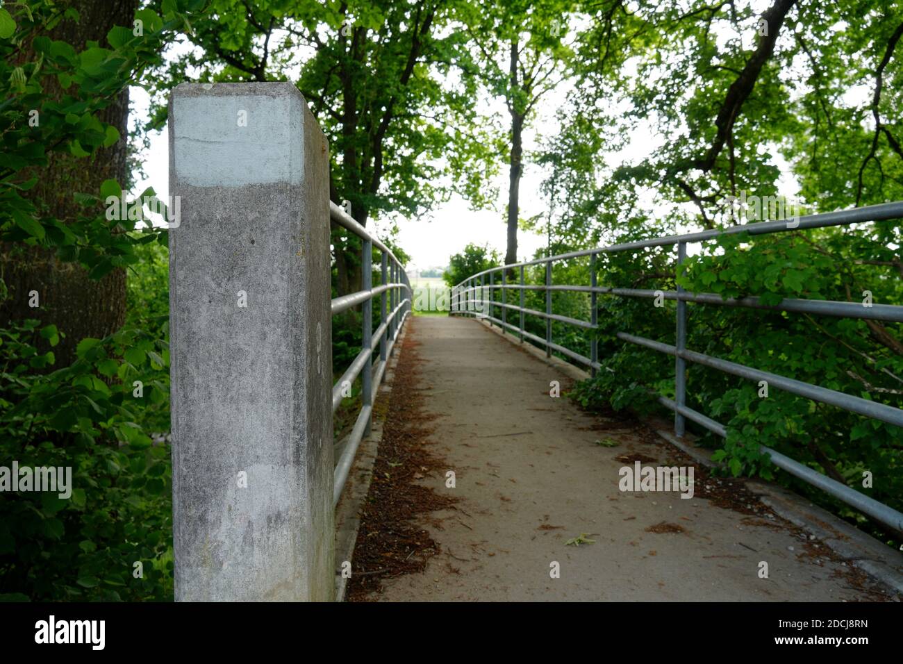 A narrow bridge with metal railings and concrete pylon surrounded by lush green vegetation diminishing in the perspective of a sun flooded land. Stock Photo