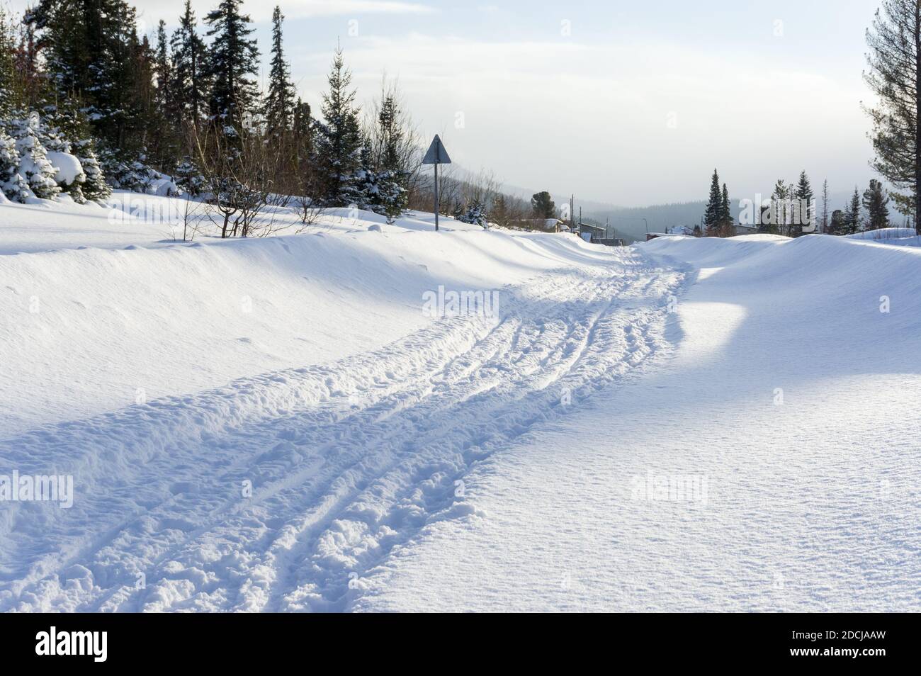 Snow-covered rural road leads to the village among the coniferous forest. Stock Photo