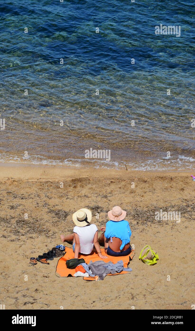 Two women sitting on their beach towels, wearing two wide-brimmed hats and watch the blue Sardinian sea Stock Photo