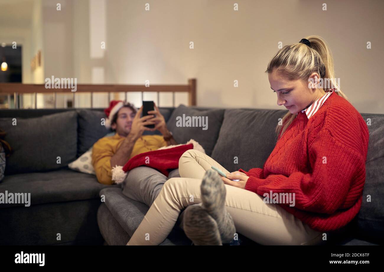 young caucasian blonde sitting on sofa with her boyfriend laying, both using their cell phones on christmas. Corona concept Stock Photo
