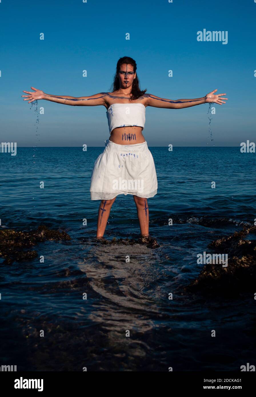 Girl with brown skin and curly hair dressed in a white cloth and her face and body painted in the sea with open arms and water falling from her hands Stock Photo