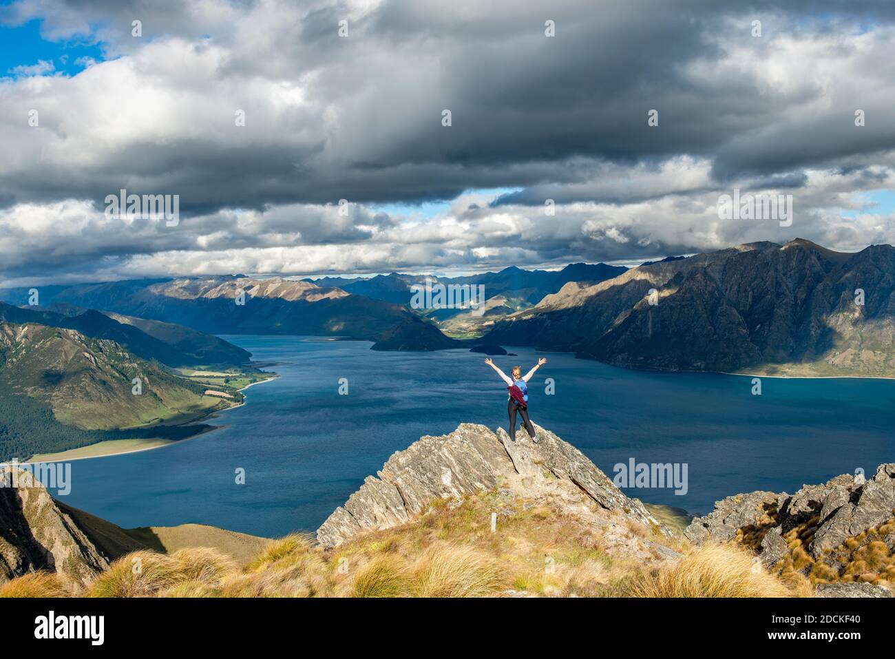 Hiker stands on a rock and stretches her arms in the air, view over Lake Hawea, lake and mountain landscape in the evening light, view from Isthmus Stock Photo