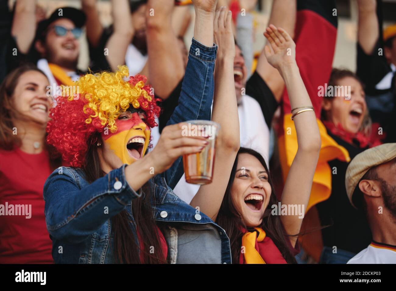 Woman in fan zone with a wig and face painted in german flag colors holding a glass of beer and cheering with crowd of spectators at sports event. Ger Stock Photo