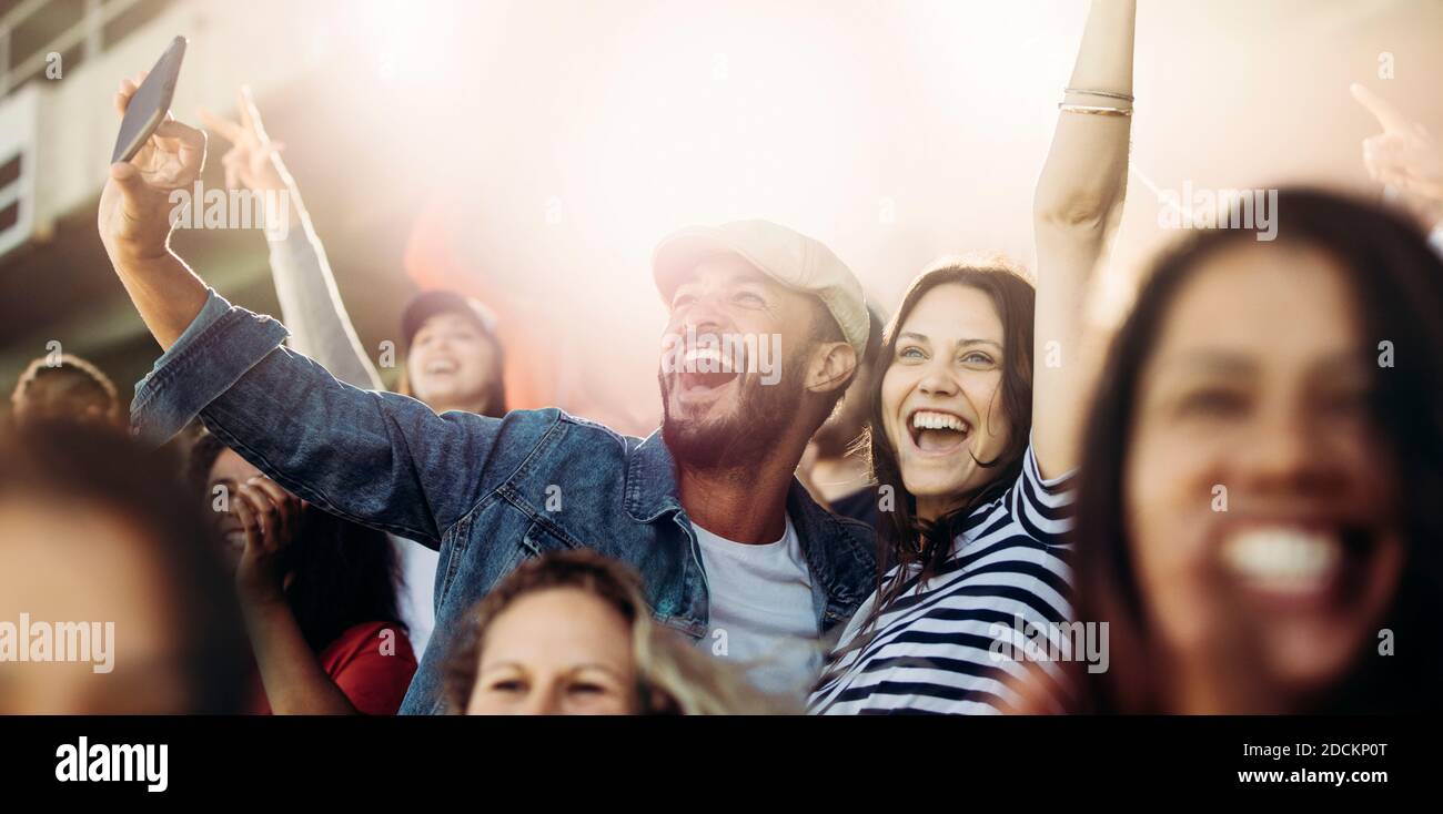 Couple taking selfie while cheering at stadium. Excited man and woman fans taking selfie while watching match in stadium. Stock Photo