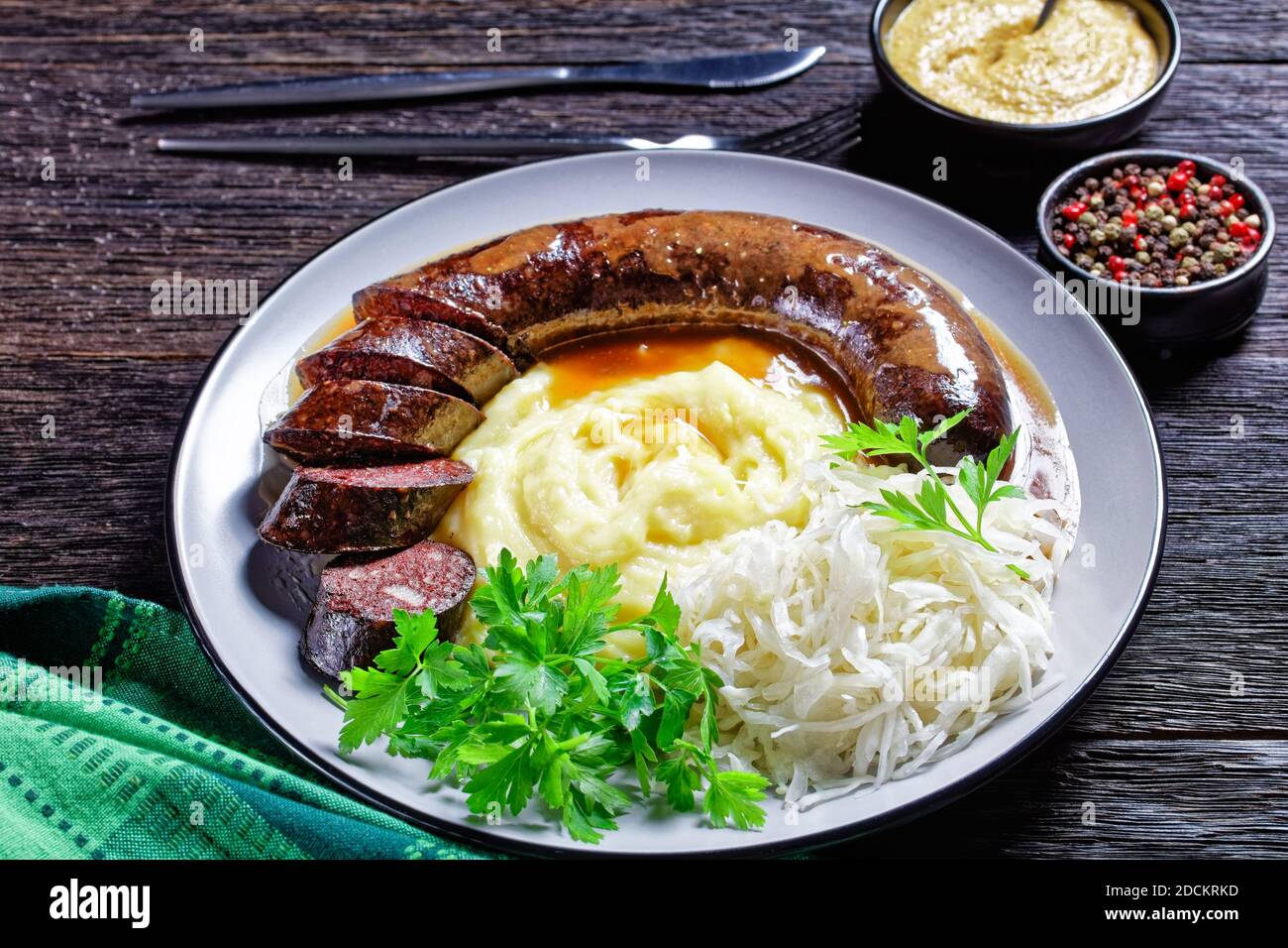 German food: blutwurst or blood sausage served on a plate with sauerkraut, mashed potato parsley, mustard, and peppercorns on a dark wooden table, top Stock Photo