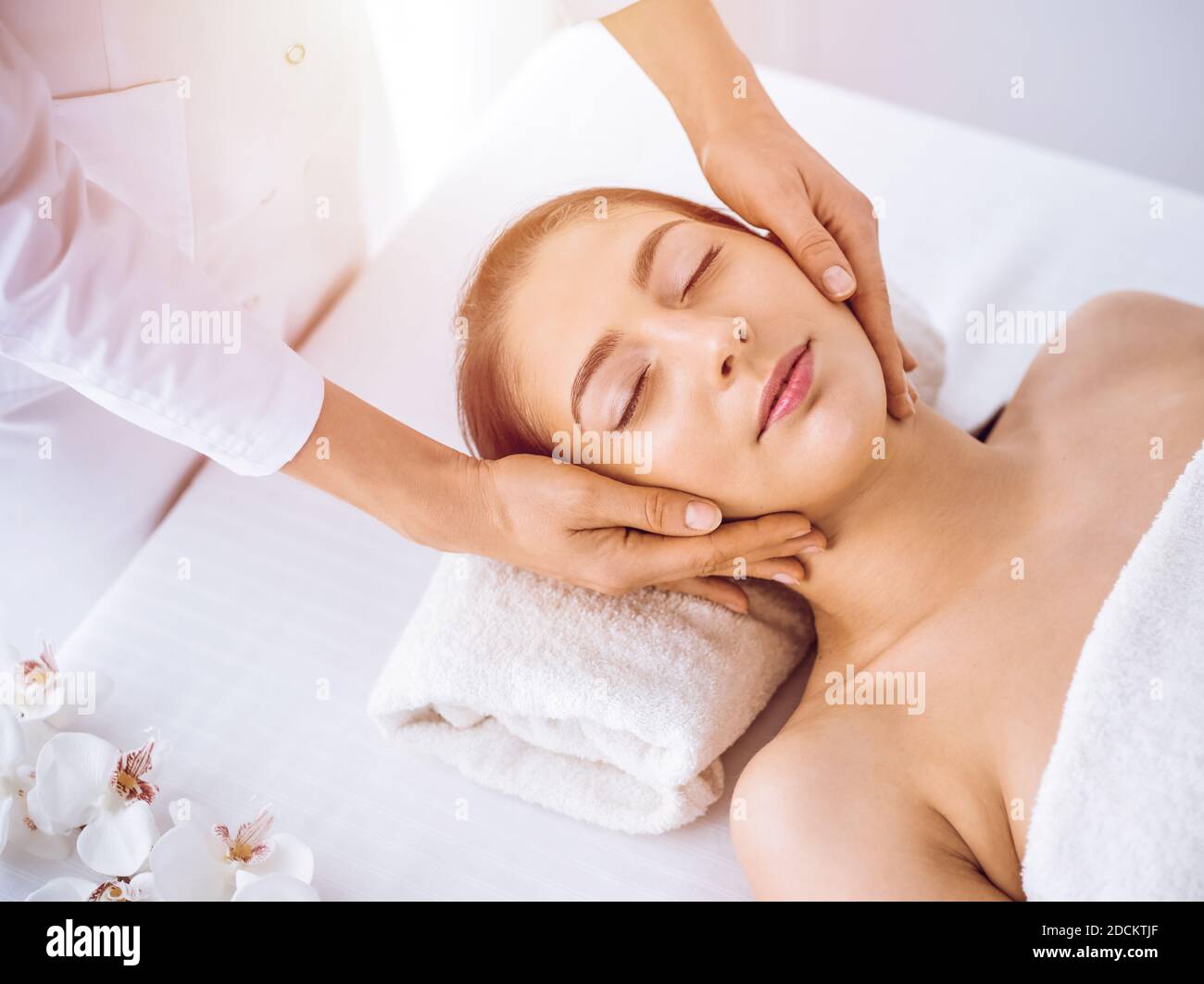 Beautiful brunette woman enjoying facial massage with closed eyes in sunny spa center . Relaxing treatment and cosmetic medicine concepts Stock Photo