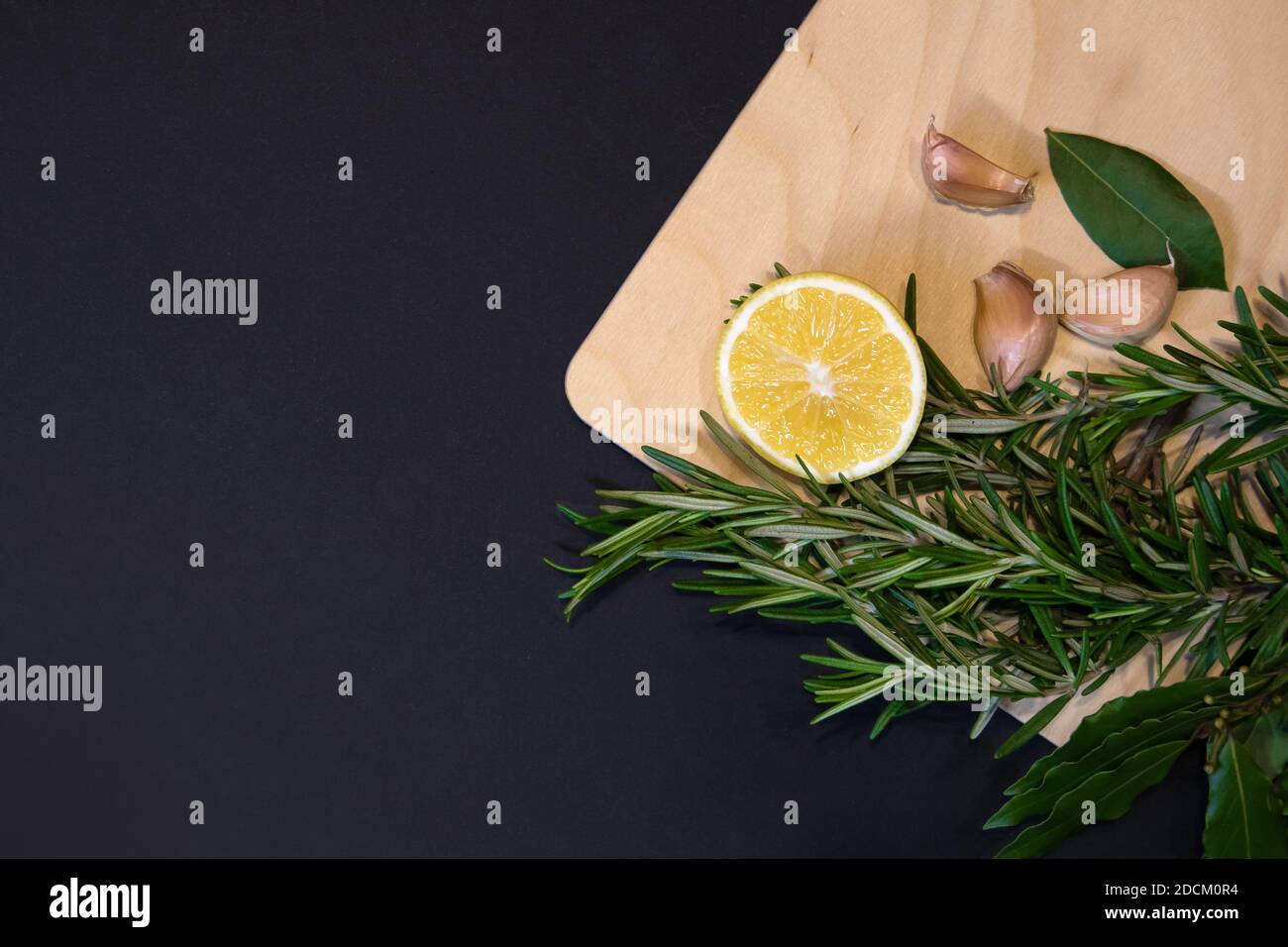 Cutting board of light wood framed with bay leaves, rosemary sprigs, lemon and cloves of garlic, designer template with dark gray background Stock Photo