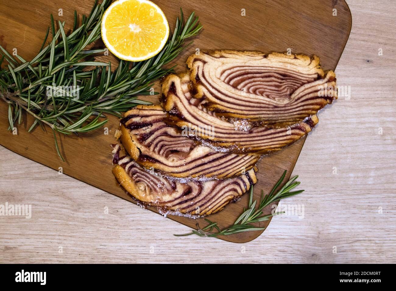 Sweet dessert served on a wooden board with sliced pastry roll and lemon half decorated with rosemary sprigs Stock Photo