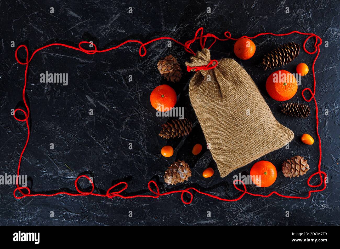 Christmas bag made of coarse burlap, cones, kumquat, tangerines or oranges, frame made of red twine on a dark background. Copy space. Stock Photo