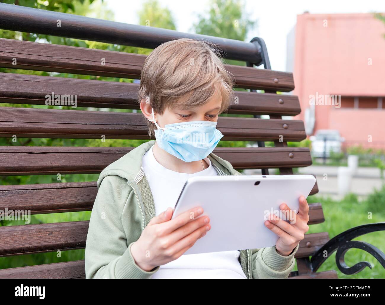 A boy with medical mask sitting on the park bench with tablet ...