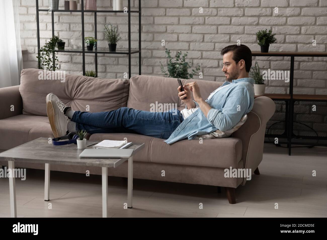 Young 30s man using mobile software application, lying on sofa. Stock Photo