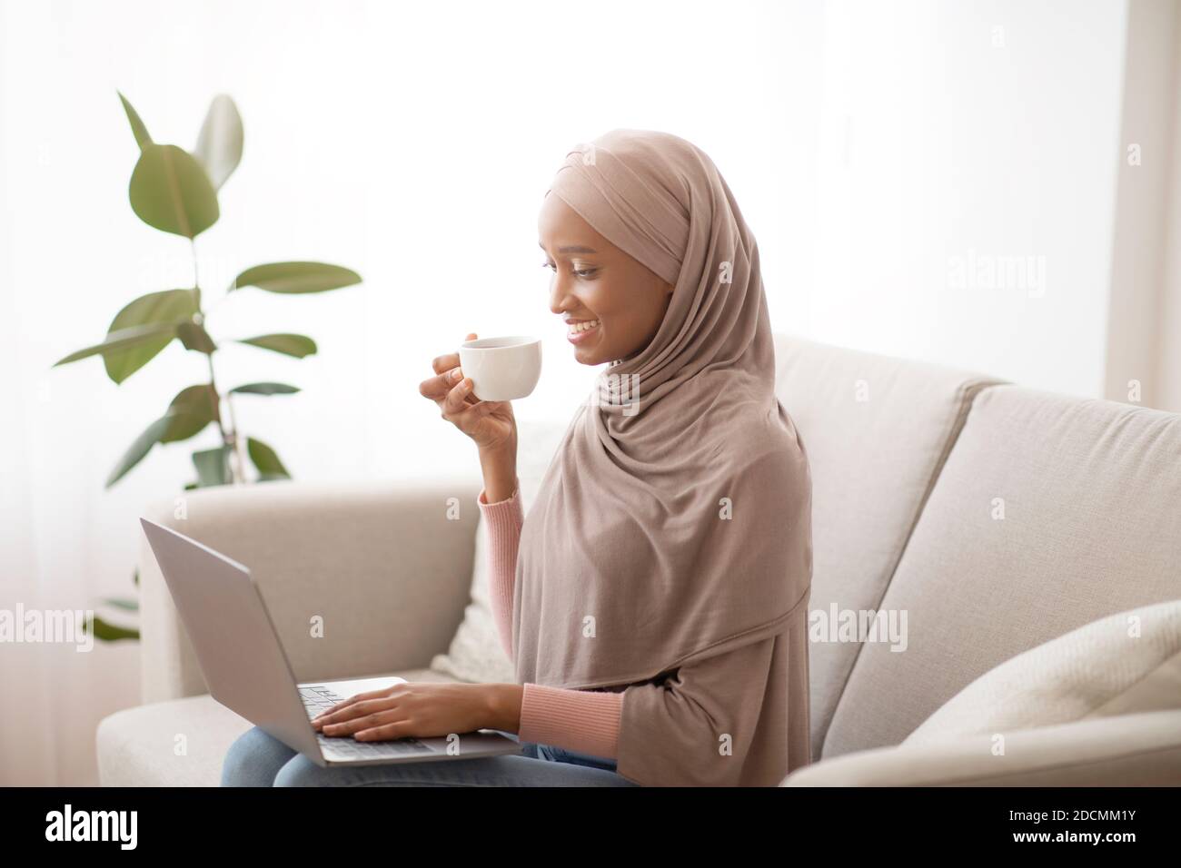 Cheerful black lady in hijab sitting on couch with laptop and drinking ...