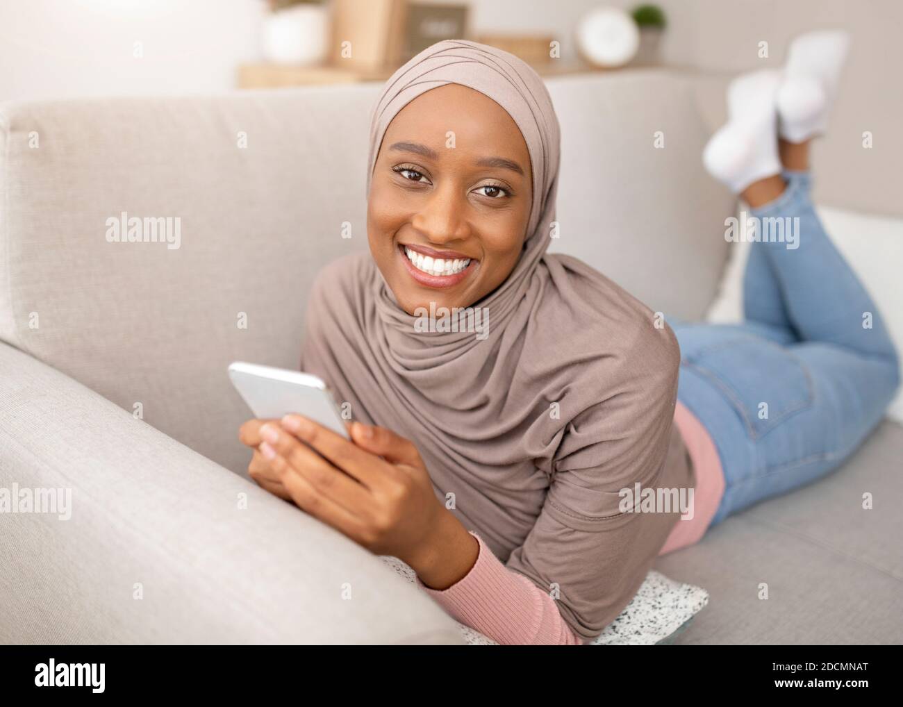 Online life. Smiling black woman in hijab lying on sofa with smartphone ...