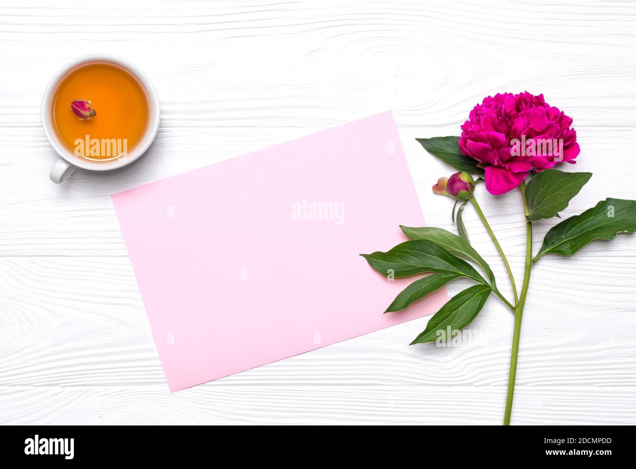 Empty paper with place for text, a cup of tee and peony flower on a white wooden background. Mockup for your design. View from above. Stock Photo