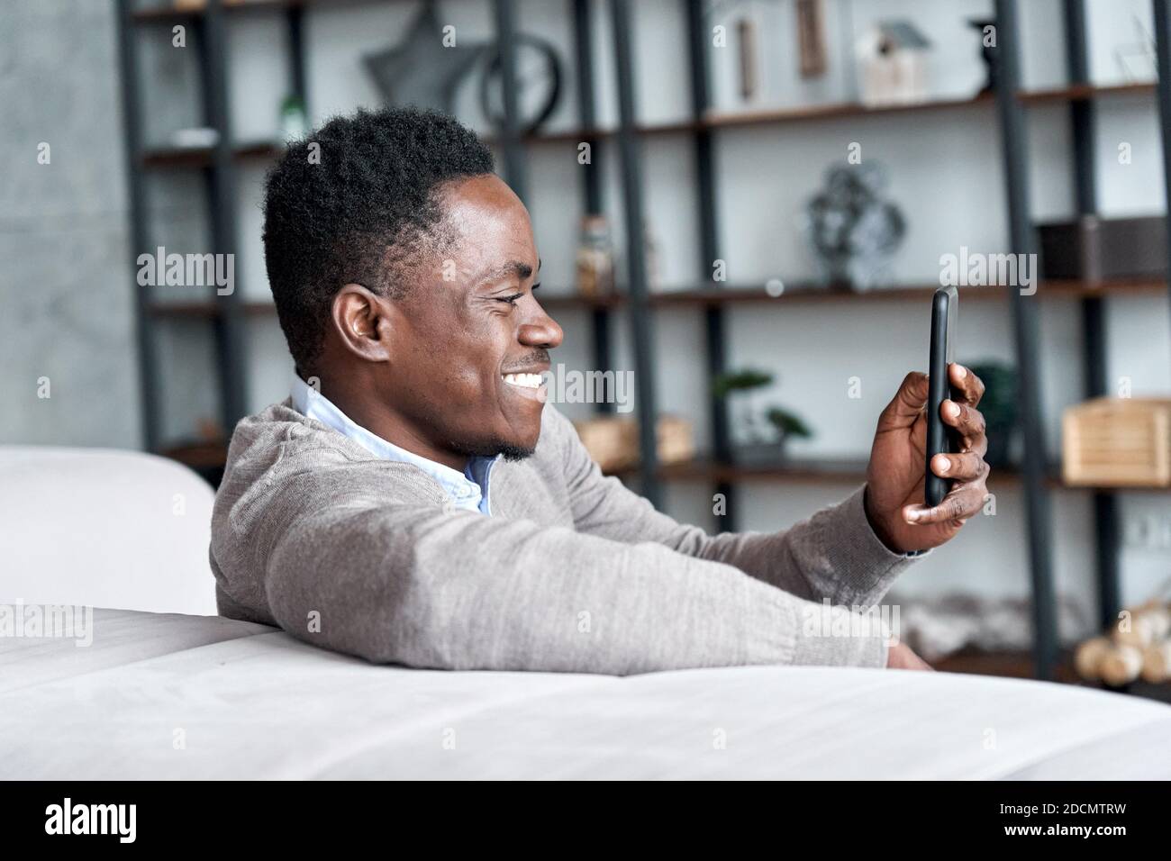 Happy african young man using mobile apps sitting on couch at home. Stock Photo