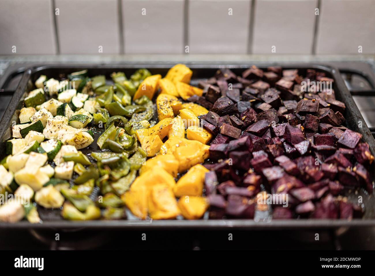 Close-up of oven-baked vegetables on black baking tray. Sliced green peperoni, yellow sweet potatoes, beetroot, green zuchinni with spice and pepper. Stock Photo