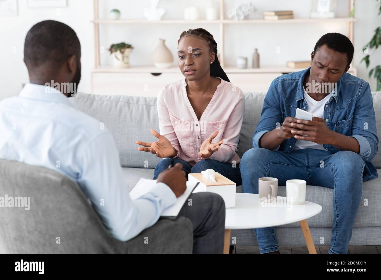 Displeased black woman blaming husband at psychologist consultation, complaining about smartphone addiction Stock Photo