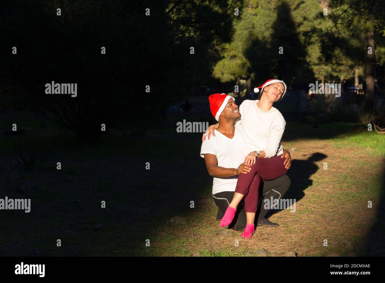 Young couple with red Christmas hats sitting on sunlight spot in forest  park. Happy woman on Brazilian man lap enjoying sun rays. Leisure time,  winter Stock Photo - Alamy