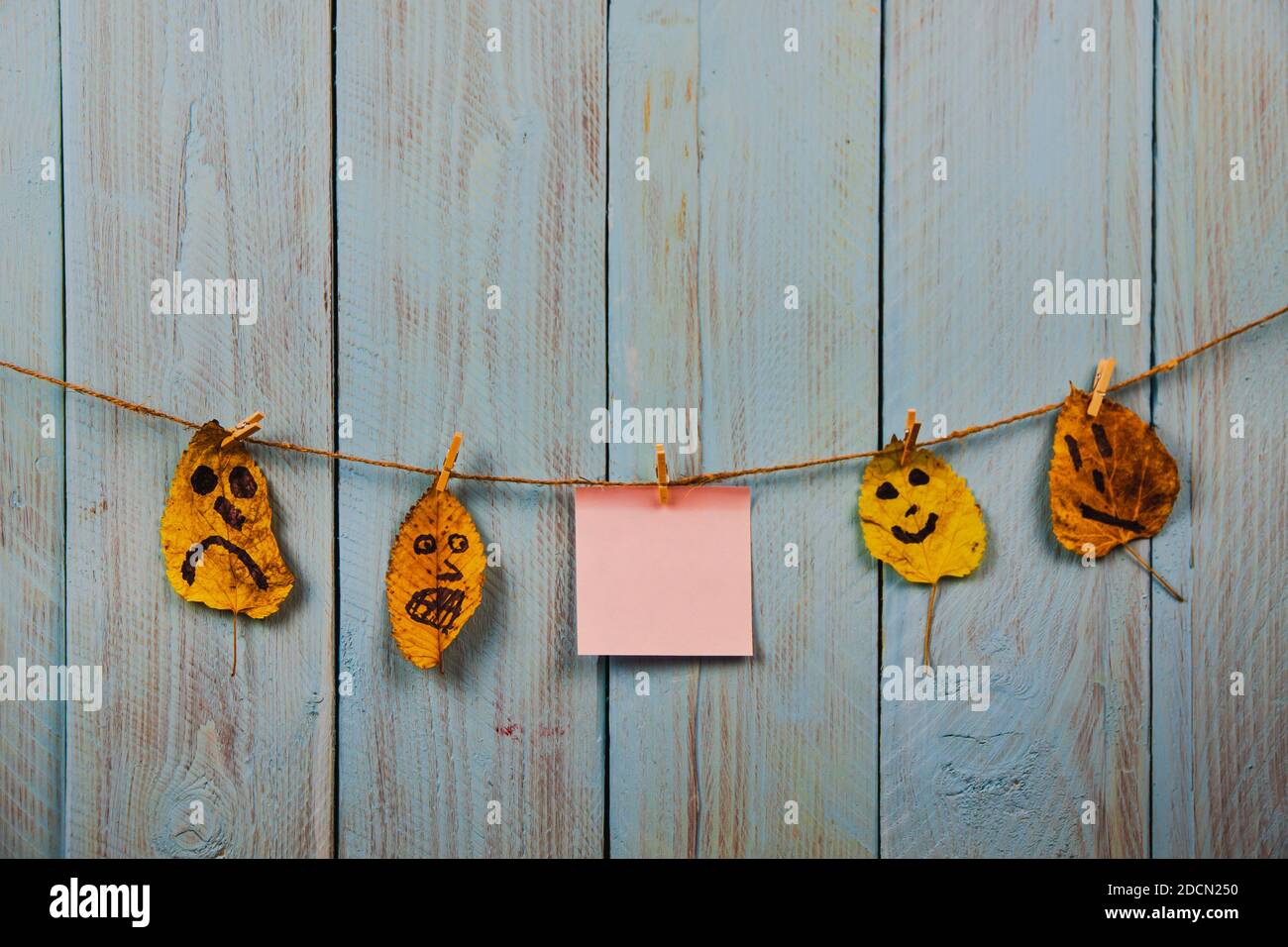 Pink note papers, and funny faces on the autumn leaves hanging on the rope against blue rustic wooden background. Copyspace. Concept of to do list Stock Photo