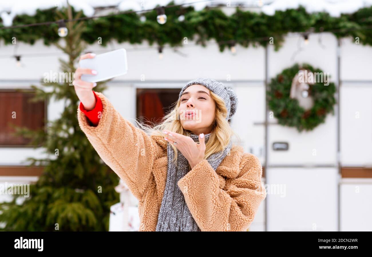 Playful Blonde Woman Taking Selfie On Smartphone Outdoors Blowing Kiss