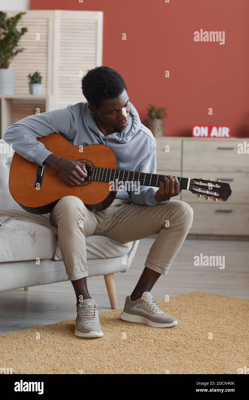 Vertical full length portrait of young African-American man playing acoustic guitar while sitting on couch at home Stock Photo